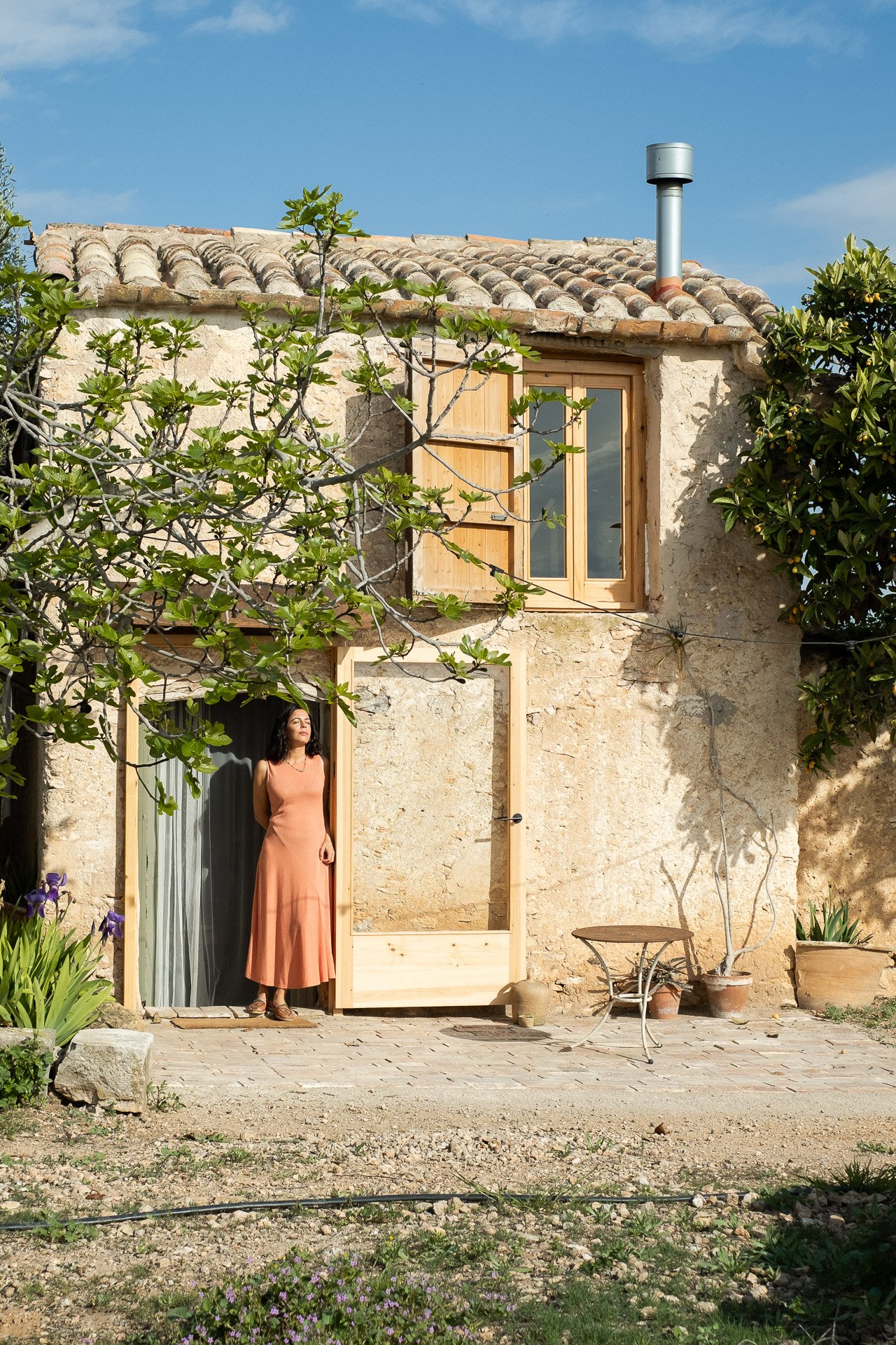 Mujer de pie en la entrada de una casa de piedra con puertas y ventanas de madera. Lleva un vestido largo color salmón y está bajo un árbol que cubre parcialmente la fachada. Hay macetas y muebles rústicos alrededor.