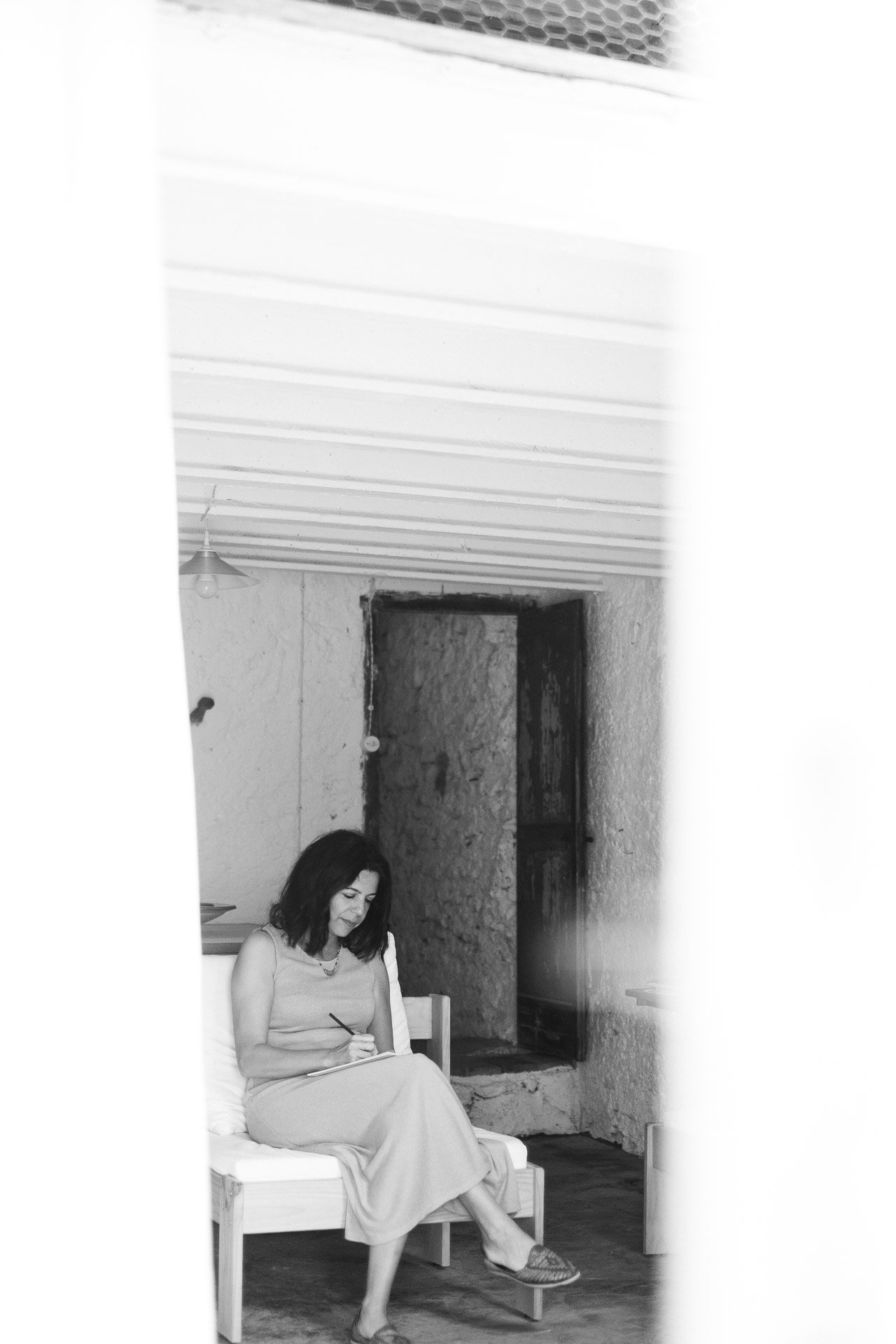 Fotografía en blanco y negro de una mujer sentada en un sofá de madera en el interior de una casa rústica. Está escribiendo o dibujando en una libreta con un bolígrafo. La habitación tiene paredes de piedra y una puerta abierta al fondo.