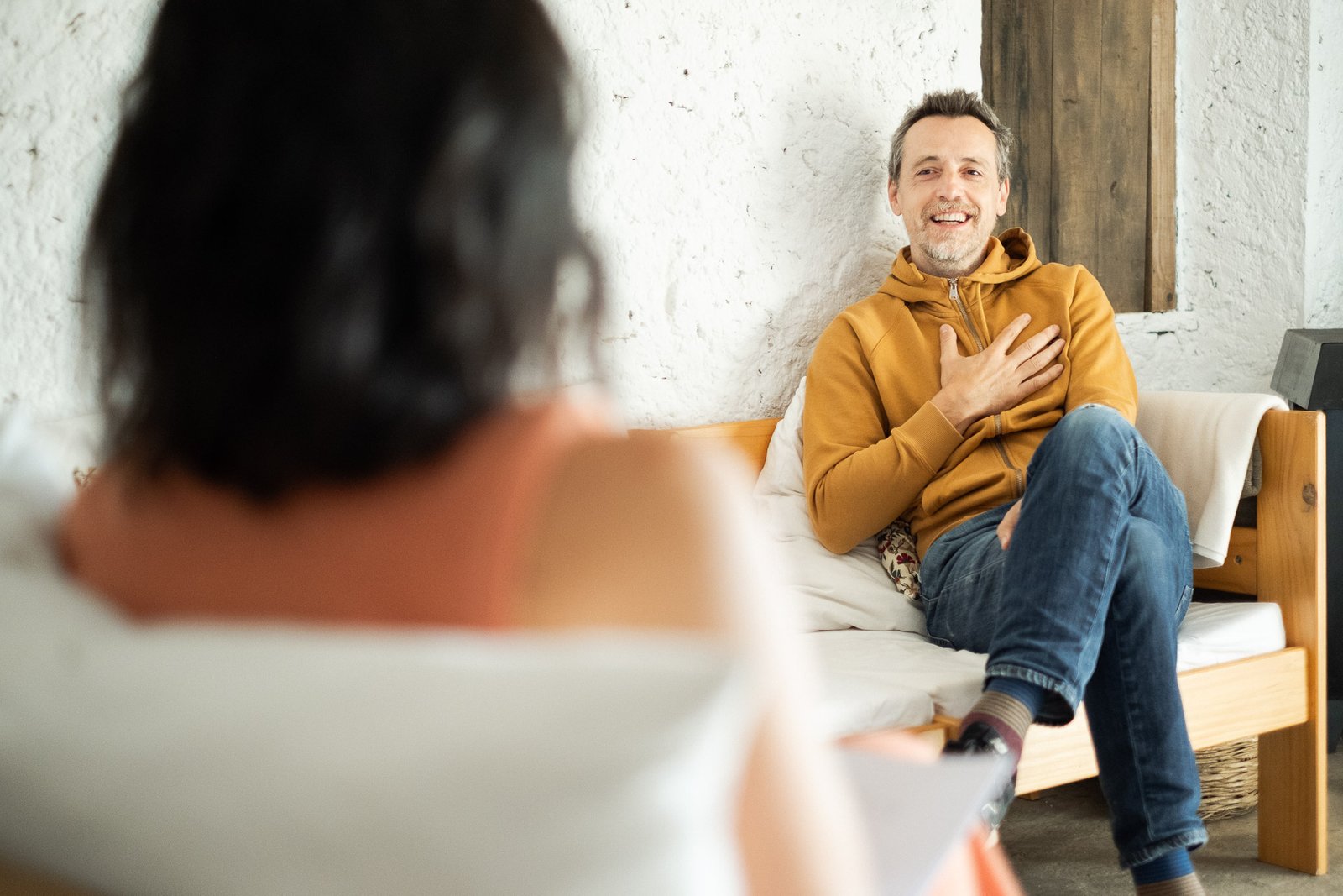 Hombre sentado en un sofá de madera, sonriendo con una mano en el pecho. Lleva una sudadera color mostaza y jeans. Frente a él, desenfocada, está una mujer que lo escucha, sentada en otro sofá.