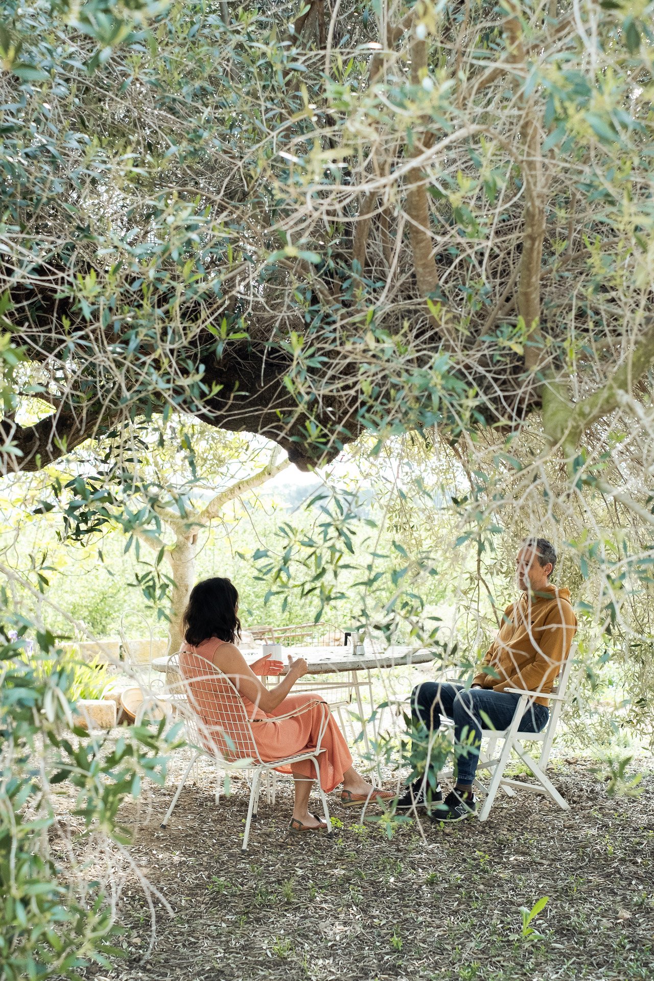 Mujer y hombre sentados en sillas blancas bajo un árbol de ramas densas. Están conversando en una mesa redonda de metal en un entorno natural, rodeados de vegetación.