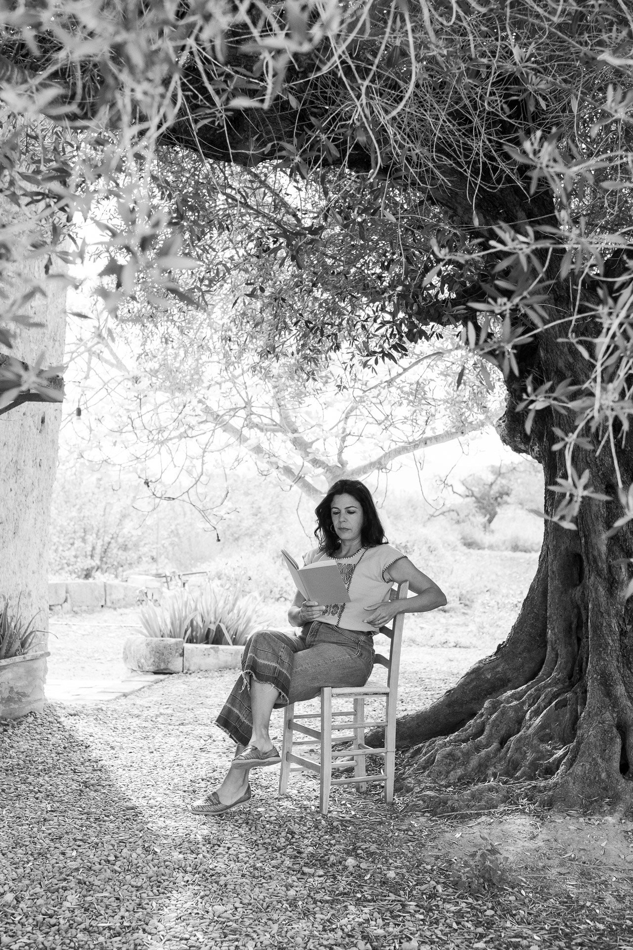 Fotografía en blanco y negro de una mujer sentada leyendo un cuaderno bajo un árbol grande. Está en una silla de madera, con pantalones vaqueros y sandalias. La escena se ubica en un entorno natural con una construcción de piedra al fondo.
